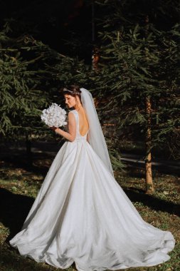 Dantel elbiseli şirin bir gelin, bir buket beyaz orkideyle parkta durarak tatilin tadını çıkarıyor. Gelinin portresi. Omuzları açık, uzun beyaz elbise.