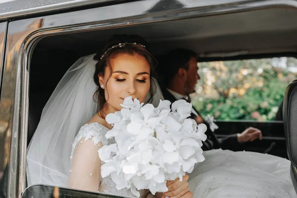 花嫁は結婚式の日に花束を持って黒い車に座っています 花嫁の肖像画 緑豊かな白いレースのドレス — ストック写真