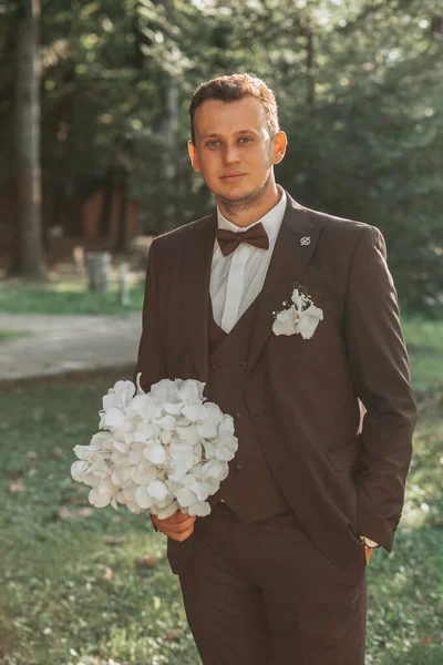 茶色のスーツに白い花の花束と蝶ネクタイを持つ森の中の若いエレガントな新郎 — ストック写真