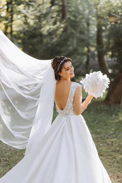 stock image A cute bride in a lace dress is enjoying the holiday, standing in the park with a bouquet of white orchids. Portrait of the bride. Long white dress with open shoulders