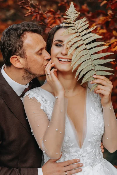 一个有着漂亮发型的女孩站在秋天的森林里 穿着一件白色的婚纱 背景是橙色的叶子 手里拿着一个蕨类枝条 她丈夫在她身后吻她 — 图库照片