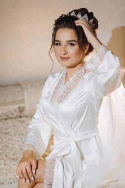 Saten bornoz giymiş esmer gelin kanepede oturur. Güzel saç ve makyaj. Düğün portresi. Samimi bir gülümseme. Gecelik giymiş güzel bir gelin poz veriyor