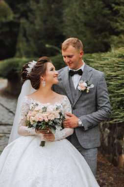 Parkta yakışıklı damat ve büyüleyici gelin birlikte. Portre. Uzun trenli lüks bir elbise. Zarif genç damat. Düğün buketi. lüks düğün