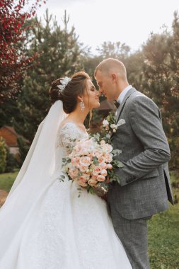 Parkta yakışıklı damat ve büyüleyici gelin birlikte. Portre. Uzun trenli lüks bir elbise. Zarif genç damat. Düğün buketi. lüks düğün