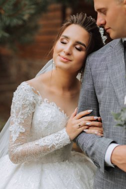 Parkta yakışıklı damat ve büyüleyici gelin birlikte. Portre. Uzun trenli lüks bir elbise. O, kolunu ona doladı. Zarif genç damat. Düğün buketi. lüks düğün