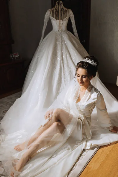 一个穿着缎子长袍的黑发新娘坐在她衣服旁边的地板上 漂亮的头发和妆容婚礼肖像 真诚的微笑 一个穿着长袍的漂亮新娘在她的衣服旁边摆姿势 — 图库照片