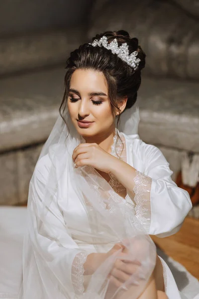 黑发新娘穿着缎子长袍 坐在沙发上 漂亮的头发和妆容婚礼肖像 真诚的微笑 一个穿着礼服的漂亮新娘正在摆姿势 — 图库照片