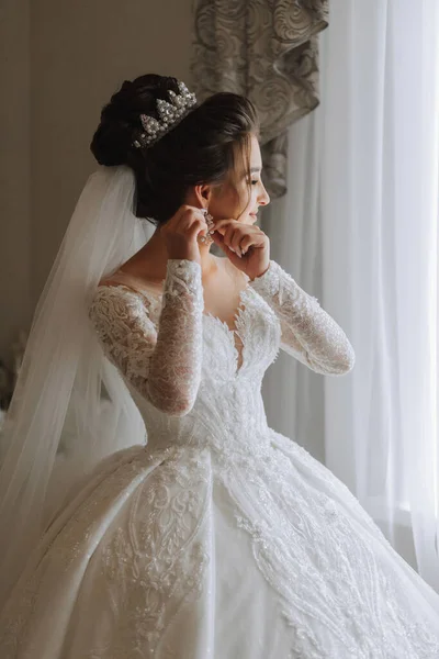 Beautiful young bride getting dressed before wedding ceremony at home. A brunette bride in a wedding dress wears earrings
