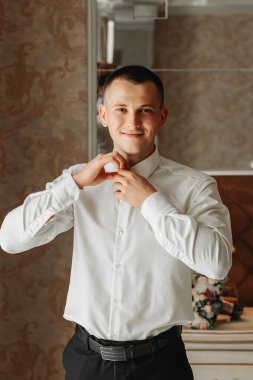 Genç, gülümseyen bir adam odasında beyaz bir gömlek ilikliyor. Damat düğün töreni için hazırlanıyor.