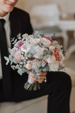 Bir otel odasında elinde bir buket çiçekle bir sandalyede oturan genç adam, siyah takım elbiseli ve kravatlı beyaz gömlekli genç bir işadamı. Damat düğün töreni için hazırlanıyor. Yakın plan fotoğraf..