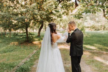 Gelin ve damat el ele tutuşur, bahçede yürürler ve birbirlerine bakarlar. Fotoğraf arkadan.