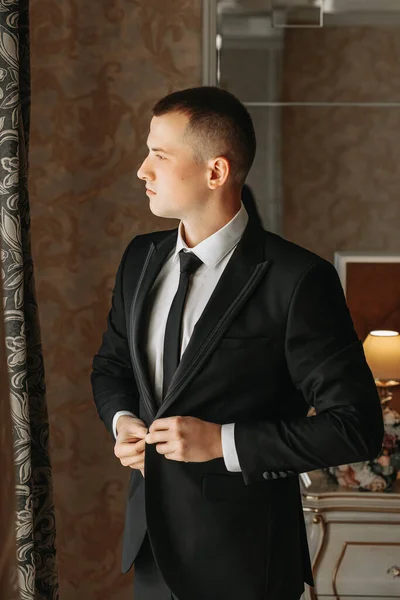 一个年轻人 一个年轻的商人 在他的房间里 在酒店房间的窗户附近 把一件夹克系在他的手上 新郎正在为婚礼做准备 — 图库照片