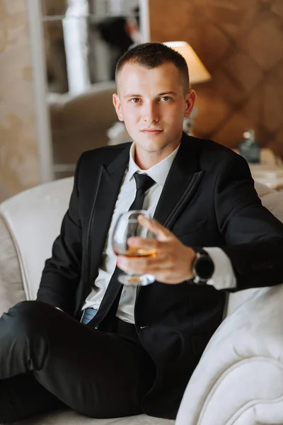 ホテルの部屋で酒を飲みながら椅子に座っている若い男黒いスーツを着た若い実業家とネクタイのついた白いシャツ 新郎は結婚式の準備をしています — ストック写真