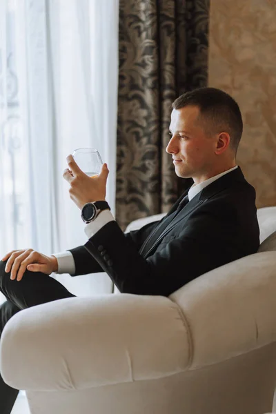 a young man sitting in a chair with a glass of alcoholic drink in a hotel room, a young businessman wearing a black suit and a white shirt with a tie. The groom is preparing for the wedding ceremony.
