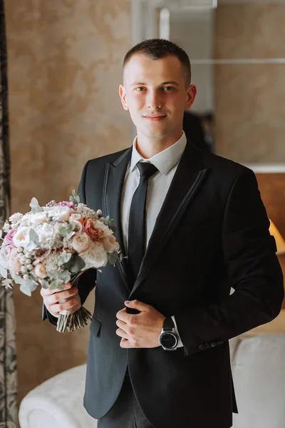 young man with a bouquet of flowers in a hotel room by the window, an adult man in a black suit and white shirt with a tie. The groom is preparing for the wedding ceremony. Close-up photo.