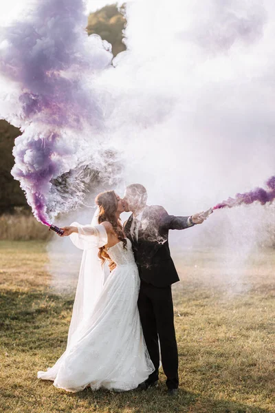 紫色の手で色の煙で遊んで花嫁と新郎は 抱擁とキス 結婚式での煙爆弾 — ストック写真
