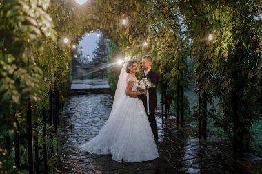 Akşam parkındaki gelin ve damat akşam ışıklarının ortasında bir buket çiçekle, doğada yeşillik. Açık havada evlenen romantik bir çift. Botanik bahçesinde düğün töreni.