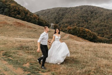 Romantik bir peri masalı çifti gün batımında ağaçlarla kaplı yüksek dağların arkasında bir tarlada koşuyor. Beyaz gelinlikli gelin, beyaz gömlekli ve siyah pantolonlu damat.