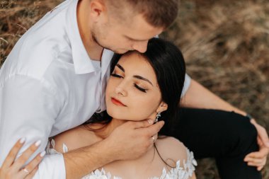 Damat ve gelin kuru çimlere uzanıp sarılır, beyaz gelinlikli kadın. Güzel bir sonbahar düğün fotoğrafı. Damat onun alnına nişan alıyor.