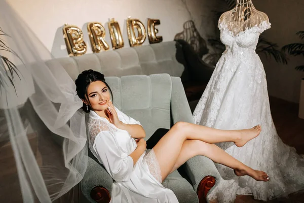 A grown-up bride sits on a chair in her room next to her wedding dress, with bare legs in a white robe. Gorgeous wedding hair and makeup