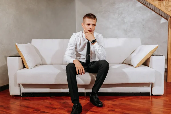 白いシャツと黒いパンツの大人の男が彼の部屋にベージュのソファの上に座っている 結婚式の準備 新郎の肖像画 フリースペース — ストック写真