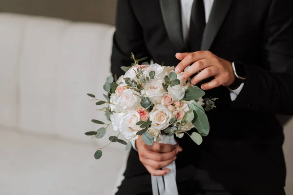 Portrait of the groom with a bouquet of flowers in his hands, sitting on the sofa in his room. Preparation of the groom for the wedding ceremony. Close-up photo