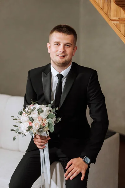 Portrait of the groom with a bouquet of flowers in his hands, sitting on the sofa in his room. Preparation of the groom for the wedding ceremony.