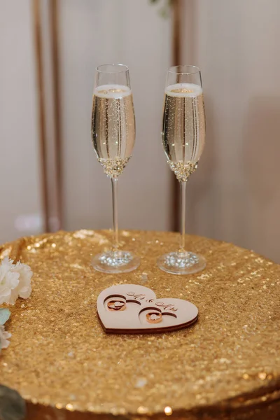 Wedding glasses filled with champagne on a table covered with gold, next to gold wedding rings on a wooden stand
