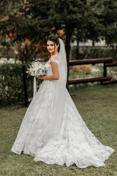 Portrait of a brunette bride in a white wedding dress with a wedding bouquet in the park. Full length photo. White lace wedding dress