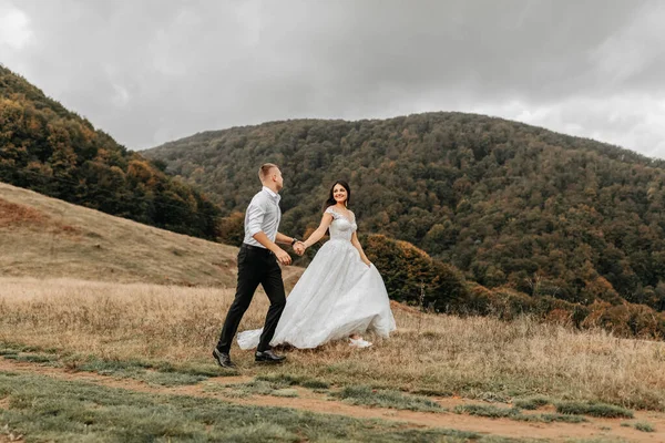 一对浪漫的童话故事 一对新婚夫妇正在落日落山后的田野里奔跑 在布满树木的高山后面 穿着白色婚纱的新娘 穿着白色衬衫和黑色裤子的新郎 — 图库照片