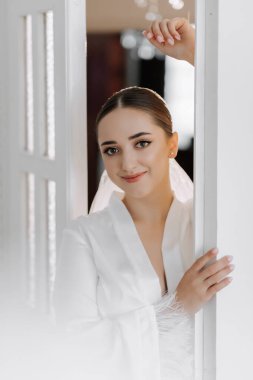 Güzel bir gelinin portresi, şık saçları ve beyaz bir bornozla balkon penceresinin yanındaki objektife bakıyor.