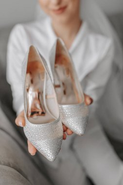 Beyaz bornozlu güzel bir gelinin portresi. Kanepede oturuyor ve yüksek topuklu ayakkabılarını elinde tutuyor.