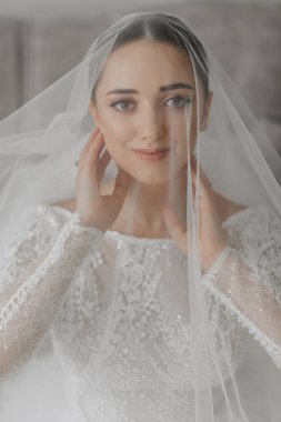 Duvağının altında saç stili ve makyajı olan güzel bir gelinin sanatsal portresi. Objektife bakar ve parmaklarıyla nazikçe yüzüne dokunur.