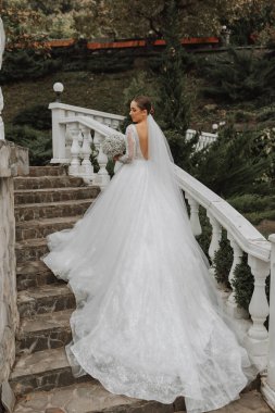 Gelinin şık bir görüntüsü, uzun trenli bir gelinlik. Elinde bir buket çiçekle gelin bahçedeki merdivenlerden tırmanıyor. Omuzlarını aç