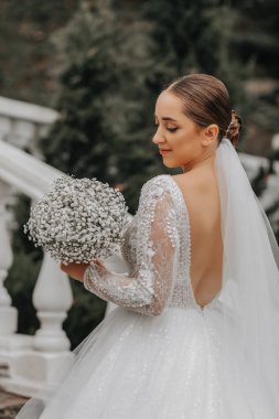 Gelinin şık bir görüntüsü, omuzları açık bir gelinlik. Elinde bir buket çiçekle gelin bahçedeki merdivenlerden tırmanıyor..