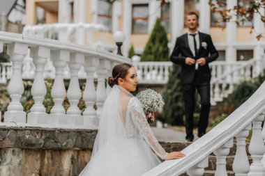 Gelinin şık bir görüntüsü, omuzları açık bir gelinlik. Elinde bir buket çiçekle gelin bahçedeki merdivenlerden damatına tırmanır..