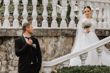 Gelin ve damadın portresi bahçede basamaklarda, damat ön planda kravatına dokunur ve karısına bakar.