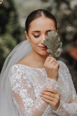 Doğada güzel bir gelin elinde meşe yaprağı, yüzünün bir kısmını kaplıyor. Genç bir gelinin yakın plan düğün portresi. Düğün fotoğrafçılığı.