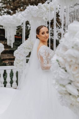 Beyaz çiçeklerle süslenmiş düğün kemerinin yanındaki parkta beyaz elbiseli tatlı esmer gelin. Yeni evlilerin düğün portresi..