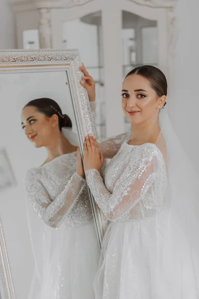 a beautiful bride in a wedding dress with closed shoulders stands by a mirror in her room, looks into the lens. reflection in the mirror