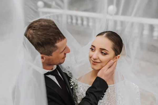 A stylish couple of European newlyweds. Smiling bride in a white dress. The groom, dressed in a classic black suit, white shirt, under a veil. Wedding in nature