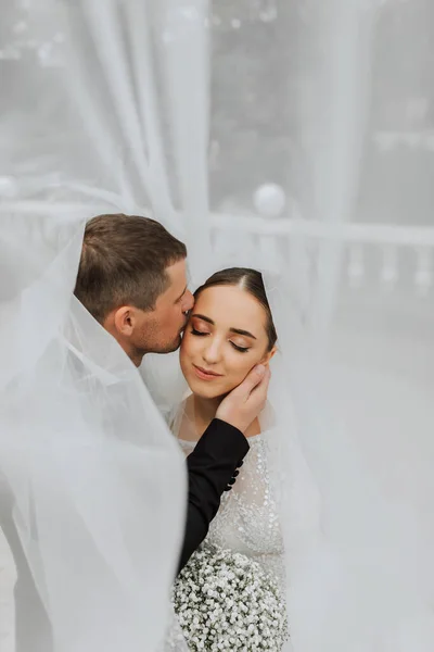 A stylish couple of European newlyweds. Smiling bride in a white dress. The groom, dressed in a classic black suit, white shirt, kisses the bride on the temple under the veil. Wedding in nature