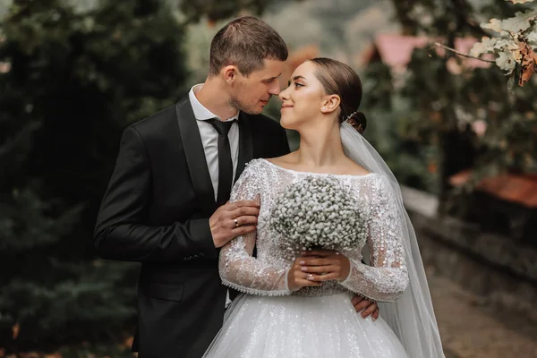 A stylish couple of European newlyweds. A smiling bride in a white dress looks at the groom. The groom, dressed in a classic black suit, white shirt, . Wedding in nature