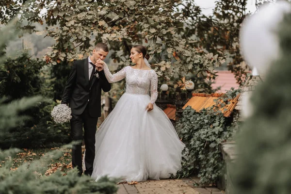 A stylish couple of European newlyweds. Smiling bride in a white dress. The groom is dressed in a classic black suit, white shirt and tie. Wedding in nature, a walk in the garden between tall trees