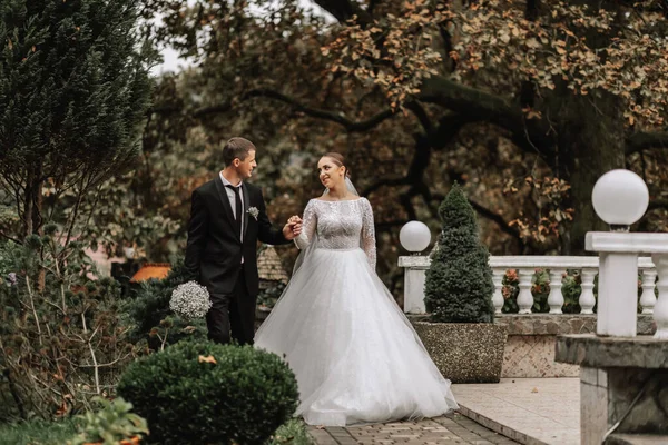 A stylish couple of European newlyweds. Smiling bride in a white dress. The groom is dressed in a classic black suit, white shirt and tie. Wedding in nature, a walk in the garden between tall trees