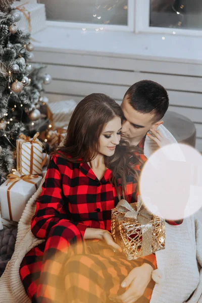 Young beautiful family couple in identical red pajamas near the Christmas tree, wrapped in a blanket, rejoice and exchange New Year\'s gifts. New Year\'s holidays and gifts under the Christmas tree
