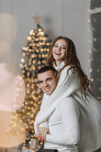 A beautiful and stylish young family in white sweaters hug and rejoice in the Christmas lights against the background of the Christmas tree. New year concept