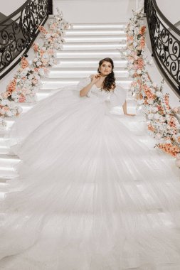 Lüks bir gelinlik içinde güzel saçlı ve makyajlı genç bir kız merdivenlerde uzanmış gül çiçeklerinin yanındaki kamera lensine bakıyor. Dikey fotoğraf