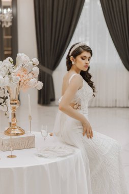 Masada elinde bir vazo çiçekle güzel bir gelin. Kadın uzun, dantelli ve arkası açık bir gelinlik giyiyor. Arkasında bir fiyonk var. Dikey fotoğraf