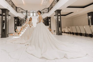Uzun trenleri olan lüks gelinlik giymiş iki güzel kız çiçeklerle süslenmiş bir restoranın merdivenlerinin yanında poz veriyorlar. Düğün salonu için fotoğraf çekimi. Şık moda.